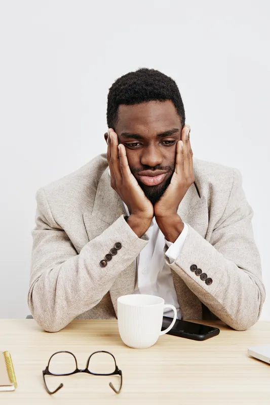 Thoughtful man sitting at desk with mug, looking worried and holding face with both hands, on plain white background, studio portrait. People emotion concept, stress, thinking, concern, loneliness, me