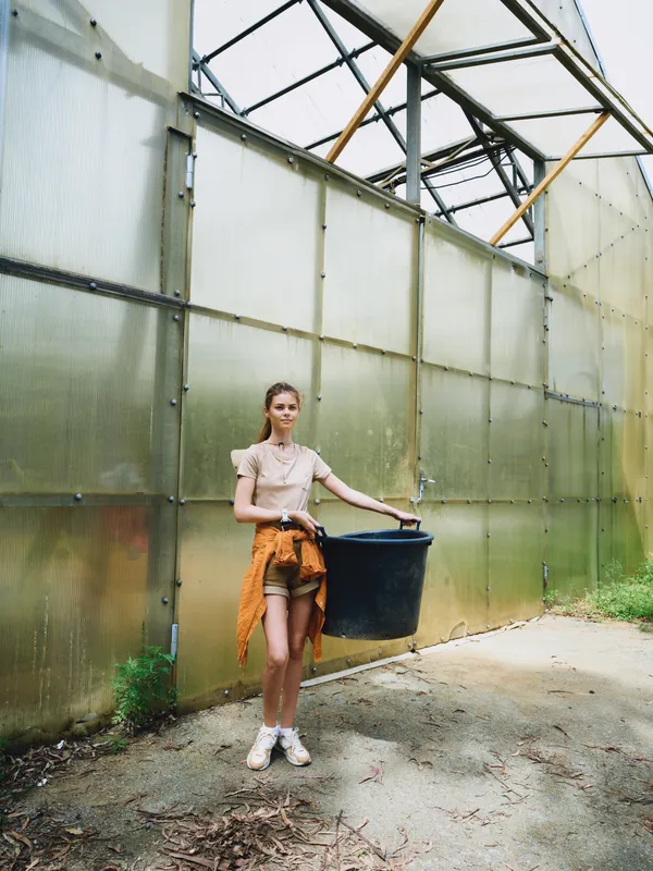 Woman greenhouse gardener carrying black bucket for gardening, casual young woman with plants on soil path, wearing shorts and sneakers inside translucent panels with sunlight.