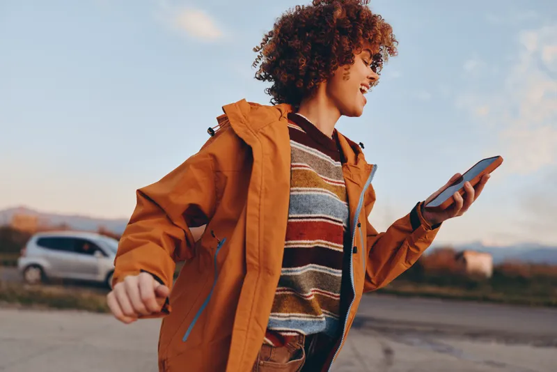 Woman lifestyle outdoors jacket smartphone walking curly hair, in orange coat checks smartphone on roadside during sunset, evening sky clouds, casual streetwear travel mood