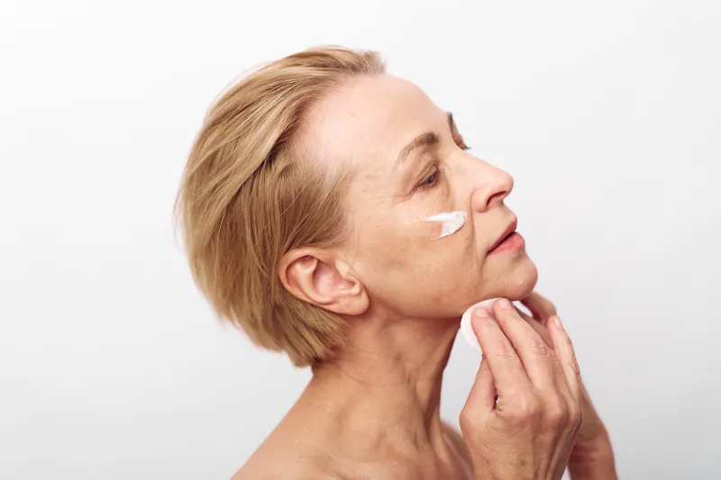 Mature woman applies white face cream on cheek with fingers, eyes closed in calm expression. Skincare and anti aging concept with soft natural light on plain white background.