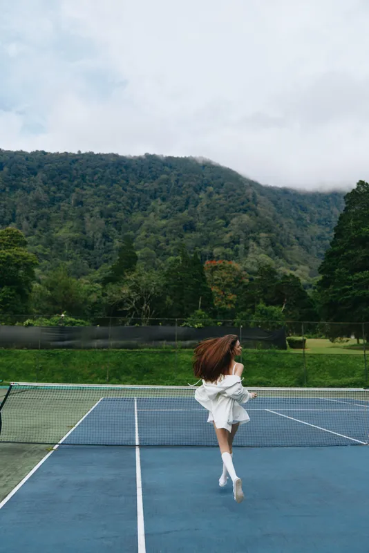 Young woman in a white dress running joyfully on a tennis court, against a backdrop of mountains and greenery, embodying freedom and adventure