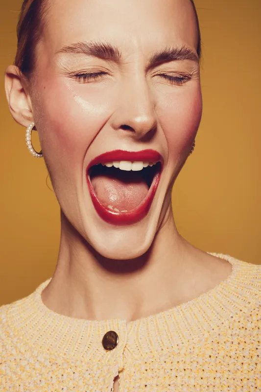 Close-up of a young woman with red lipstick winking and laughing. She wears a yellow sweater and hoop earrings against a warm brown background. Concept of joy and beauty.