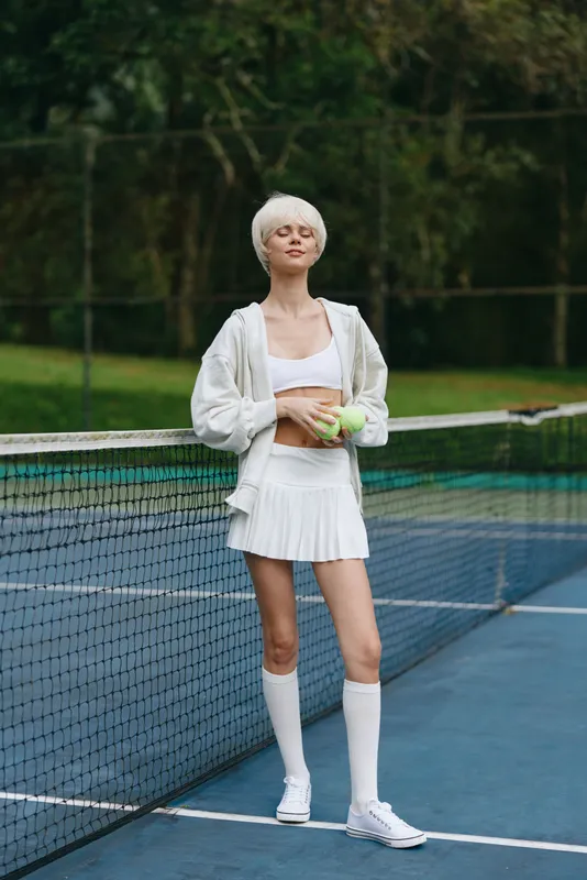 Young female tennis player in white sportswear, smiling and holding a tennis ball on a sunny court, showcasing an energetic and healthy lifestyle