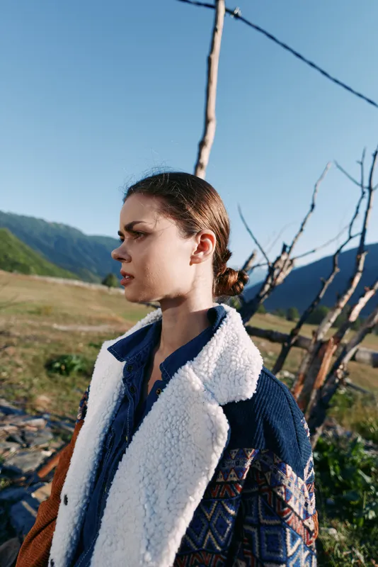 Woman portrait outdoors in mountains wearing patterned jacket with shearling collar, profile view in countryside field. Natural light, thoughtful gaze and rustic landscape for travel and lifestyle.