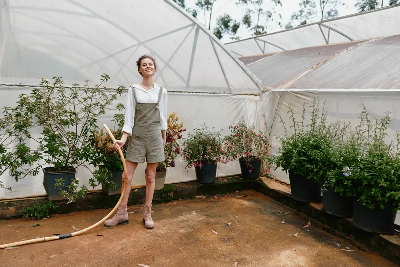 Young woman in a greenhouse wearing overalls and smiling while holding plants, surrounded by greenery, showcasing a joyful gardening concept in a natural setting