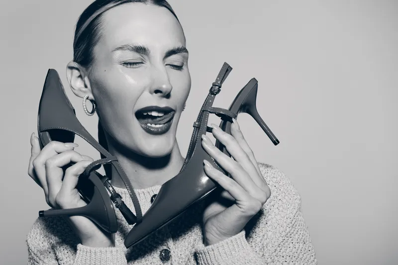 Young white woman with fair skin holds two high heel shoes near face with playful expression and closed eyes. Concept of fashion, footwear, joy, and personal style.