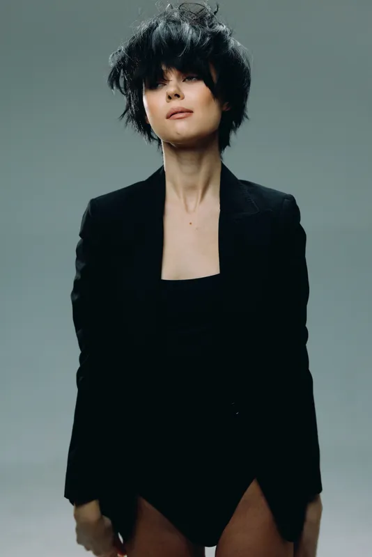 Stylish woman with short black hair wearing a black blazer against a neutral background, exuding modern confidence and minimalistic elegance.
