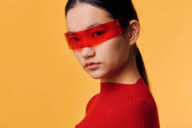 Young woman wearing red stylish transparent glasses and red top looking confidently at camera with serious expression against orange background. Portrait with modern fashion and minimalism style.