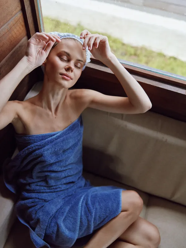 Relaxed woman in blue towel and hair wrap enjoying calm moment by window, natural light highlighting peaceful face and soft skin texture indoors spa setting.