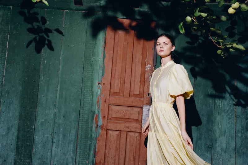 Stylish woman in a pale yellow dress stands by a wooden door on a teal wall, creating a serene fashion portrait with soft natural light and shade.