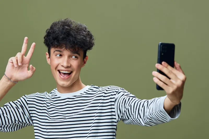 Young man taking a selfie with a smartphone, smiling happily and making a peace sign, wearing a striped shirt against an olive green background.