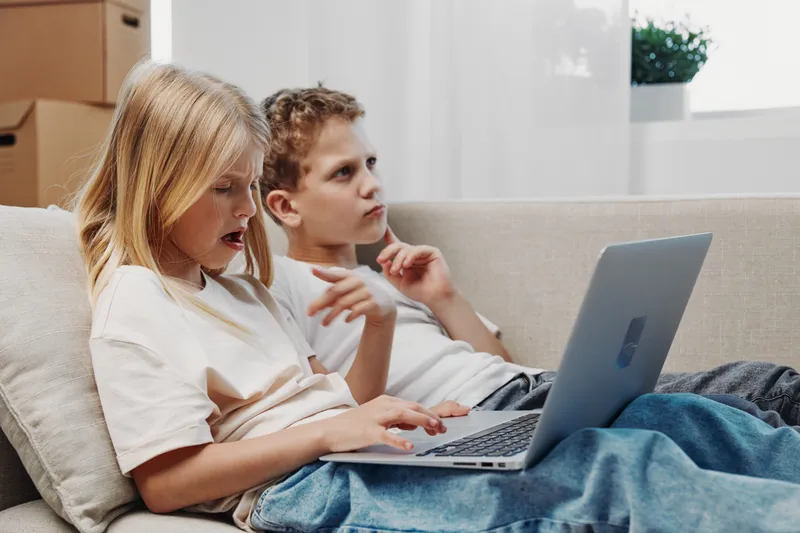 Two focused kids sitting on sofa using laptop together, learning or playing in cozy home environment with natural light.