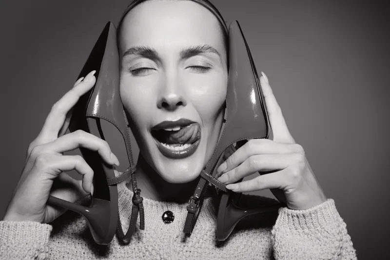 Young white woman with pale skin holds red high heel shoes near face with eyes closed and tongue out in black and white studio portrait. Concept for fashion and emotions.