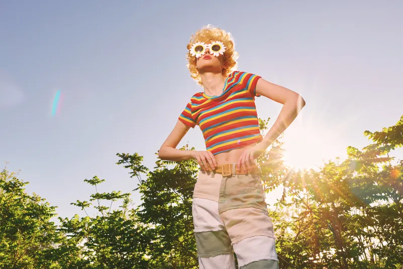 A woman stands outdoors under bright sun wearing a colorful striped crop top and patchwork pants, accessorized with floral sunglasses, posing with playful summer mood and carefree vibe sun