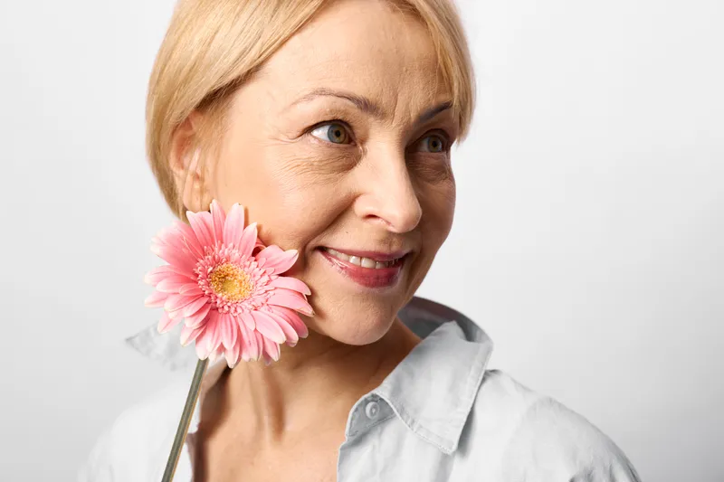Smiling woman portrait with pink flower closeup, blonde mature face, light studio background, beauty and skincare concept, wellness mood, gentle eye contact, soft light, spring