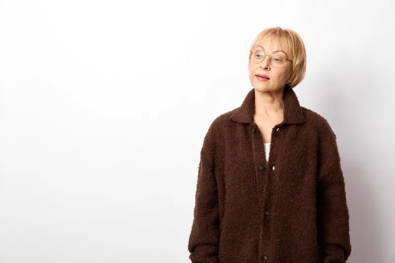 Woman older glasses coat portrait indoor on white background, looking to side with calm expression, warm brown outerwear, soft studio light, minimal space today