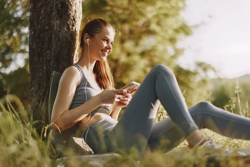 Young woman enjoying music outdoors in activewear, surrounded by greenery, smiling and relaxed Perfect for concepts of wellness, fitness, and outdoor activities