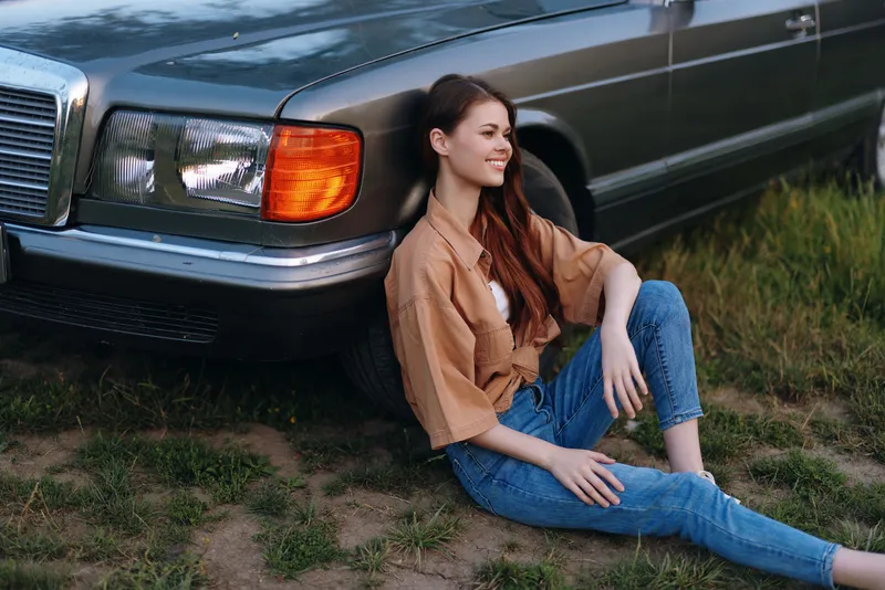 Young woman with long hair in casual outfit sitting by vintage car in grassy field, smiling and enjoying sunny day, evoking a feeling of freedom and nostalgia.