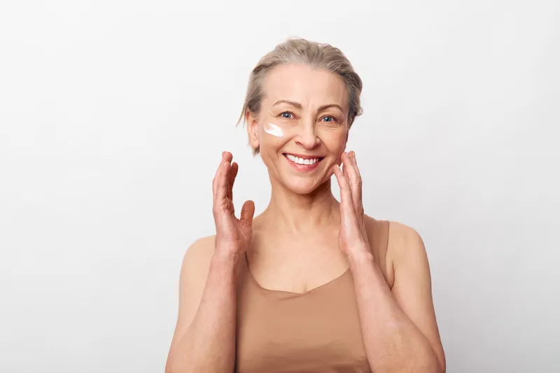 Smiling mature woman applies moisturizer cream on her face with hands, showing natural beauty and skincare routine on white background. Concept for aging skin care and wellness.
