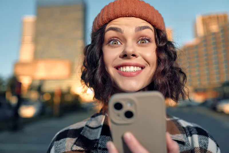 Woman with smile holding smartphone in city street wearing beanie during golden hour, candid lifestyle portrait that evokes authenticity, mindful living and emotional storytelling glow.