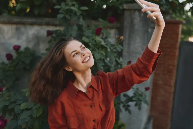 Smiling woman with long hair taking a selfie in a red shirt outdoors, garden setting, casual lifestyle moment, self expression and cheerful mood for social media