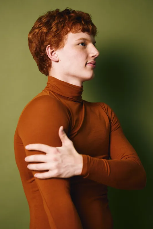 Young man with red hair wearing a brown turtleneck sweater posing in profile against green background with arms crossed showing thoughtful and calm expression.