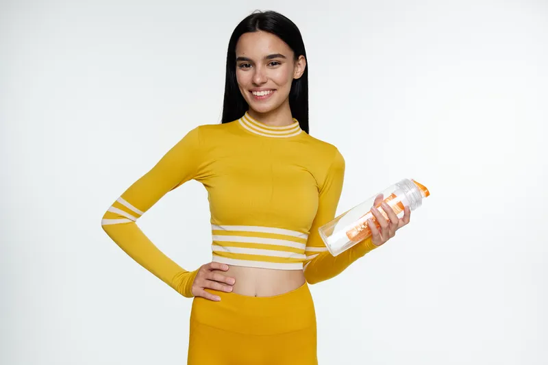 Woman sport fitness posing with a water bottle, wearing yellow activewear, smiling and confident on a bright white background, healthy lifestyle concept.