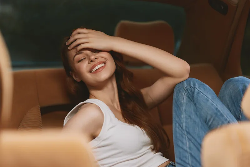 Young woman smiling joyfully in a car, wearing casual outfit, showcasing carefree emotions, warm tones, and a relaxed ambiance.
