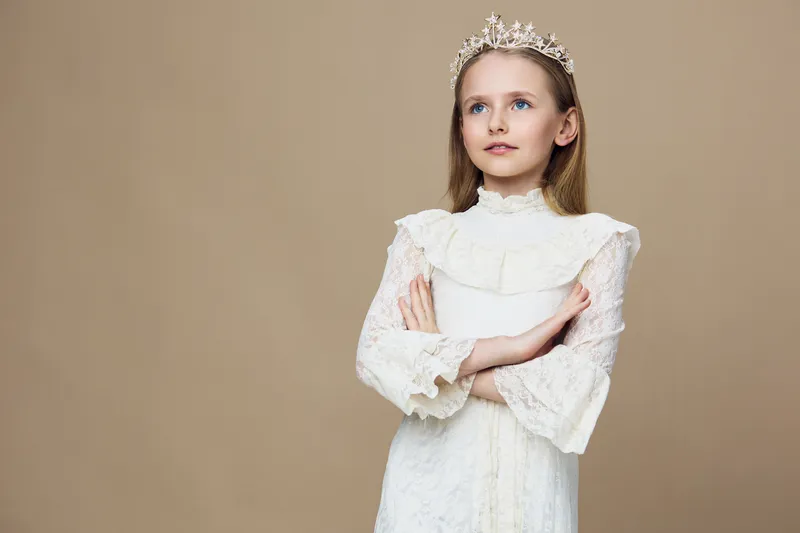 Elegant young girl in a crown wearing a white dress, with long hair and a confident pose against a soft beige background, embodying royalty and innocence