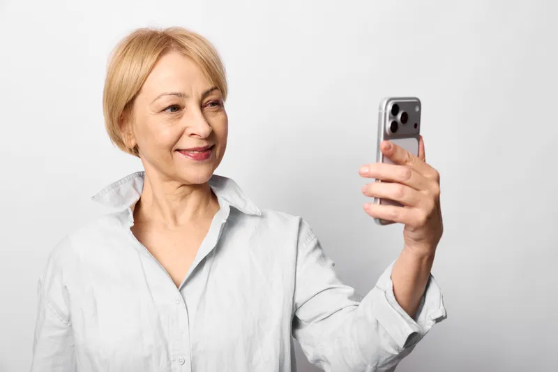 Elderly woman takes selfie with smartphone, smiling portrait, holding phone near face, casual light shirt, neutral studio background, copy space for social media profile