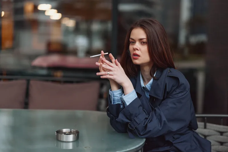 A young woman sits at an outdoor cafe, cigarette in hand, moody gaze, urban backdrop, cool tones, relaxed yet tense moment