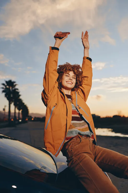 Woman lifestyle outdoor sunset scene, smile on her face, curly hair, jacket and shorts, leaning on car hood with arms raised, warm golden light, coastal road