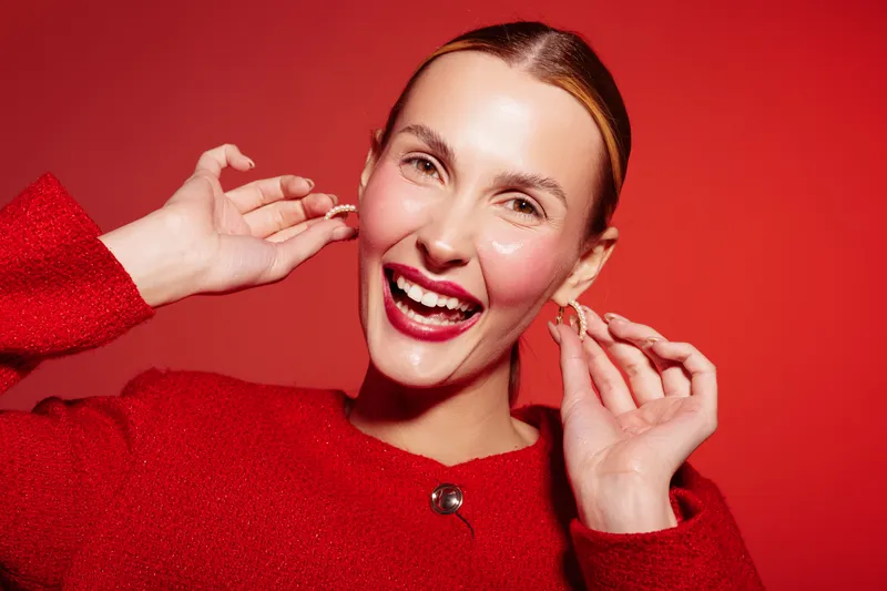 Happy woman in red sweater smiles brightly while adjusting earrings against red background. Close-up portrait with natural makeup and joyful expression. Concept of fashion and beauty.