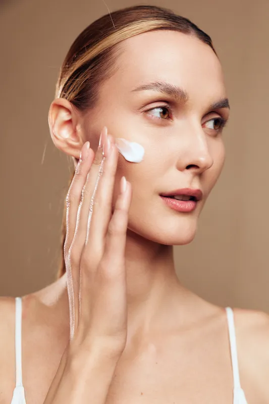 A young woman with clear skin applies white cream on her cheek using her fingers. The close-up shot highlights skincare routine and natural beauty concepts indoors.