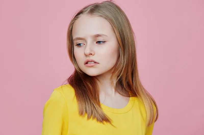 A young girl with light brown hair wearing a yellow top, looking thoughtfully to the side against a soft pink background, evoking a sense of innocence and contemplation