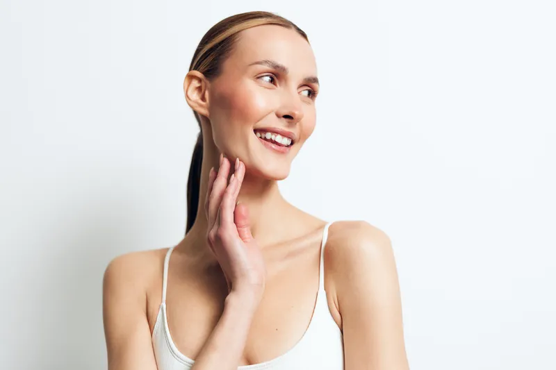 Happy young woman with clear skin and natural makeup smiles gently while touching her neck, posing in white top against plain white background with positive expression.