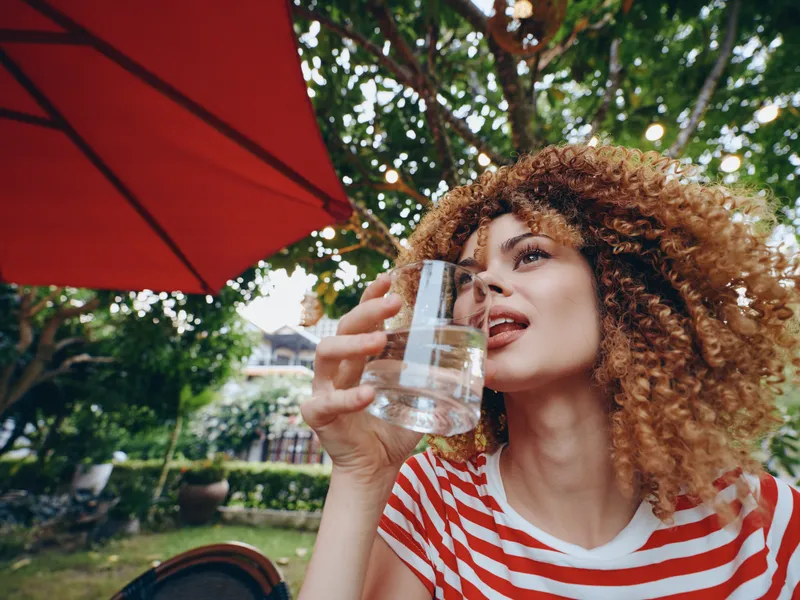 Portrait of a woman with curly hair sipping water from a glass outdoors, wearing a red striped shirt, captured in a sunlit garden cafe setting.