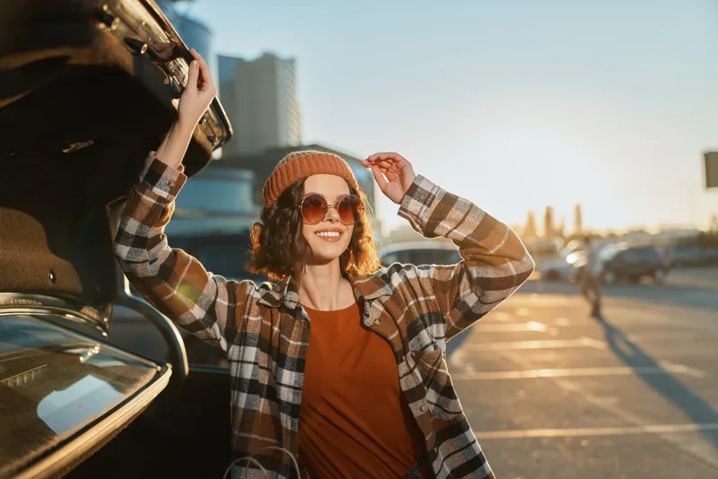 Woman with sunglasses and beanie by car trunk in parking lot at sunset, plaid jacket and warm smile, candid authenticity, golden hour glow and emotional storytelling vibe.