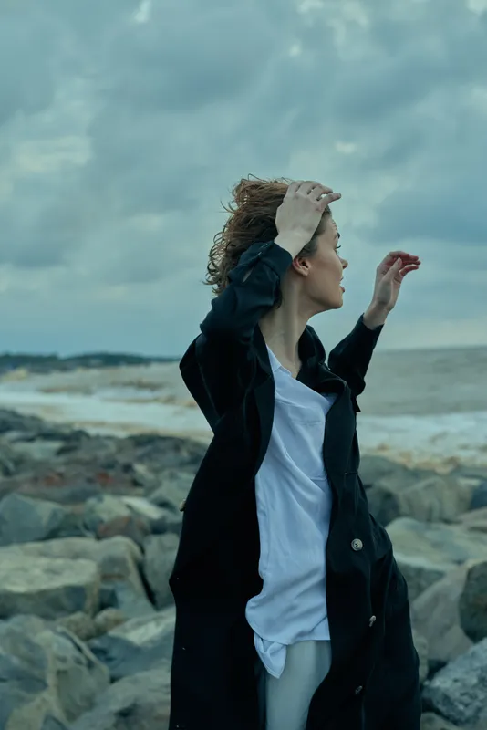 Windy coastal scene with a woman on a rocky shore, jacket over a white shirt, wind in hair, moody ocean backdrop and dramatic cloudy sky