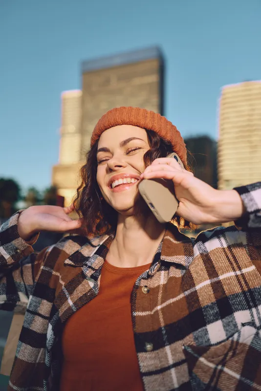 Woman smile on phone in city at golden hour, candid moment of authenticity and joyful expression with beanie and plaid jacket, mindful living and emotional storytelling in urban portrait.