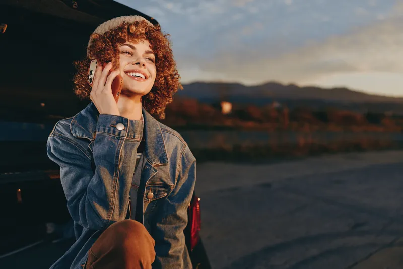 Woman lifestyle outdoors, smiling in denim jacket with curly hair, touching cheek, sunlit sunset sky, calm roadside with distant mountains, warm golden hour, candid portrait