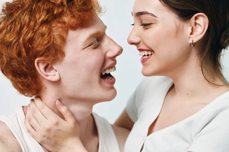 Young couple smiling and enjoying close embrace indoors. Man and woman with happy expression showing affection and connection in casual clothing on white background.