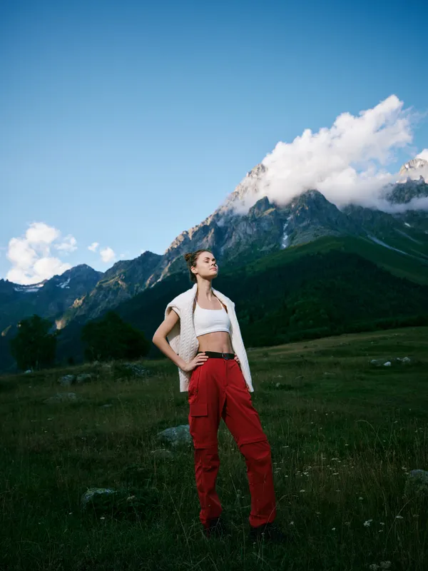 Mountain landscape with a woman in red cargo pants and white top, standing in open meadow, clear sky, conveying adventure, exploration, and outdoor style