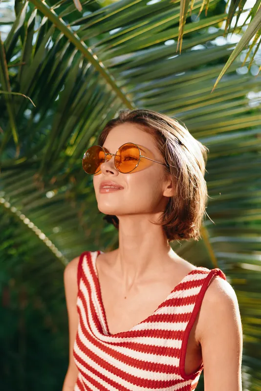 A stylish woman wears a red and white striped top and round sunglasses, gazing upward under lush palm leaves, creating a bright, tropical outdoor mood.
