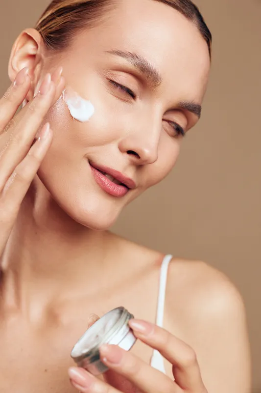 Close up of a woman applying moisturizer on her cheek, soft glow, calm expression, skincare routine in progress, creamy texture, isolated on neutral background