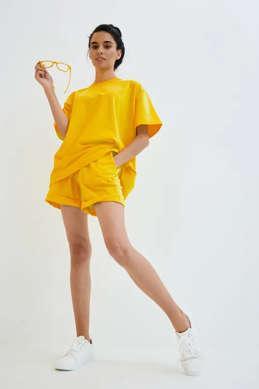 Young woman in bright yellow casual outfit with shorts and oversized t-shirt, standing confidently against white background, holding yellow glasses, sporty and relaxed style.