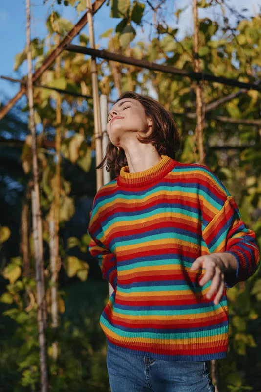 Young woman in a colorful striped sweater stands in an outdoor garden with short hair, sunlight on her face, relaxed pose and peaceful expression, enjoying a cozy casual moment.
