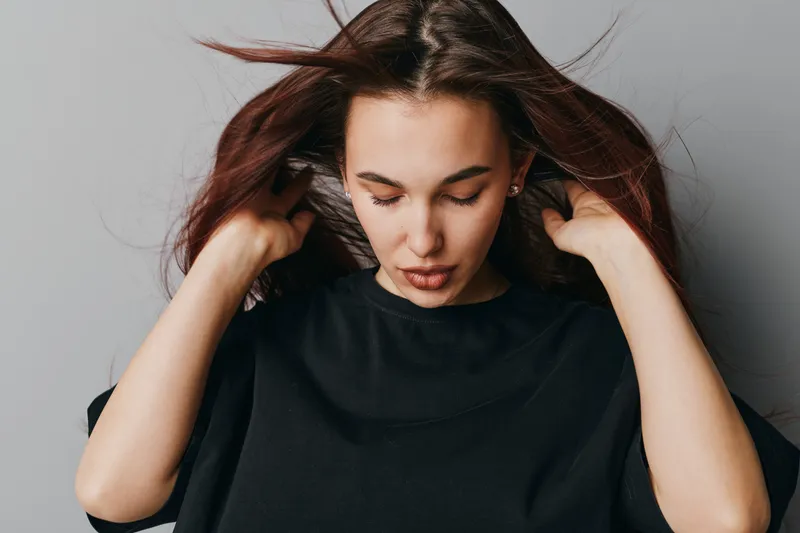 Young woman with long hair wearing black shirt looking down with hands near head against gray background. Casual style, thoughtful expression and natural makeup in studio portrait.