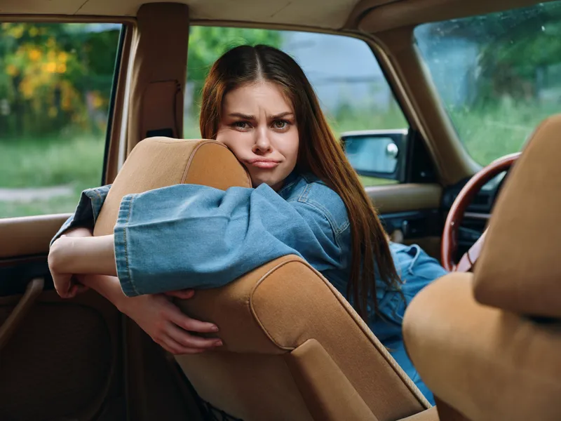 Young woman showing frustration while sitting in a vintage car, wearing a denim jacket, amidst a natural outdoor backdrop rich in greenery.