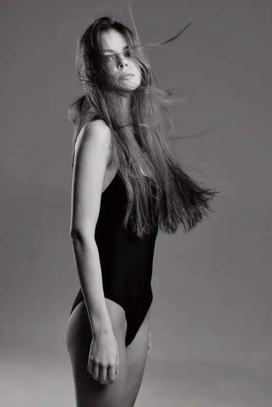 black and white portrait of young woman with long hair in simple black bodysuit, calm expression, studio background